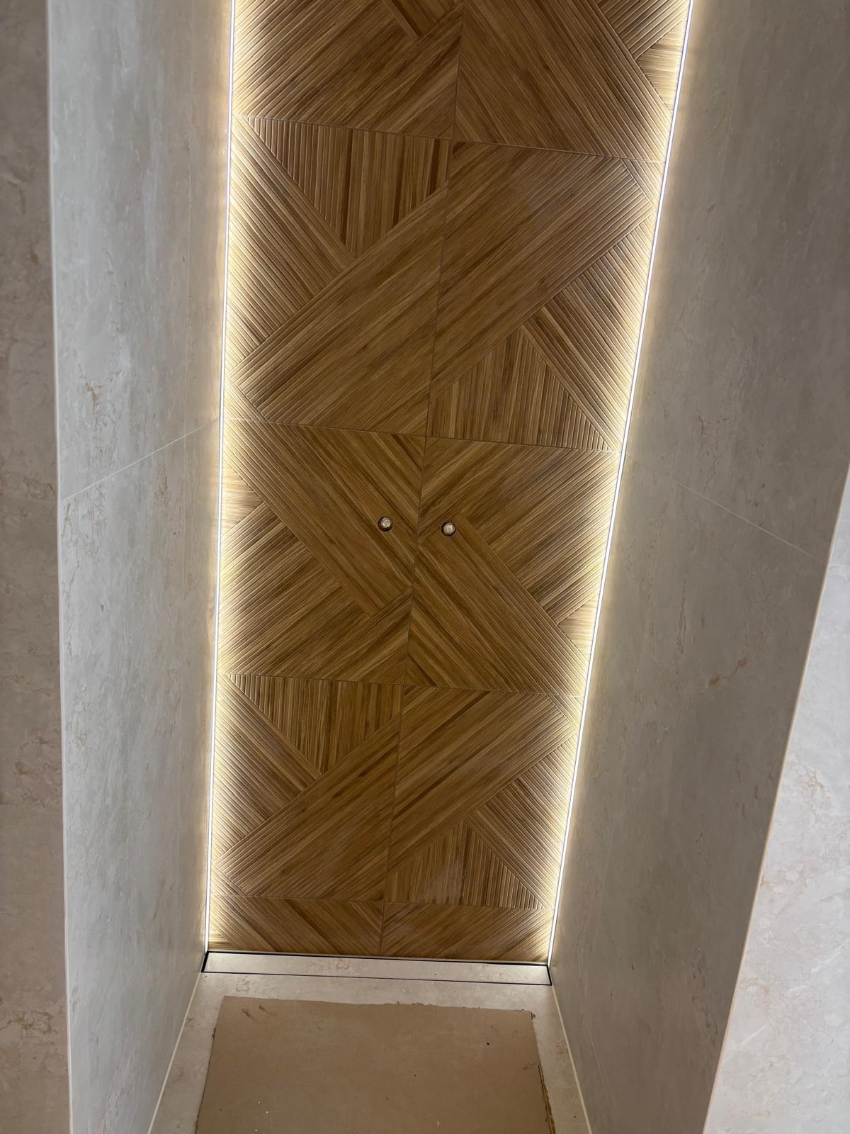 Nowoczesna łazienka z drewnopodobną, geometryczną okładziną ścienną, podświetloną listwami LED. Jasne płytki na ścianach i podłodze. Widoczne punkty świetlne.