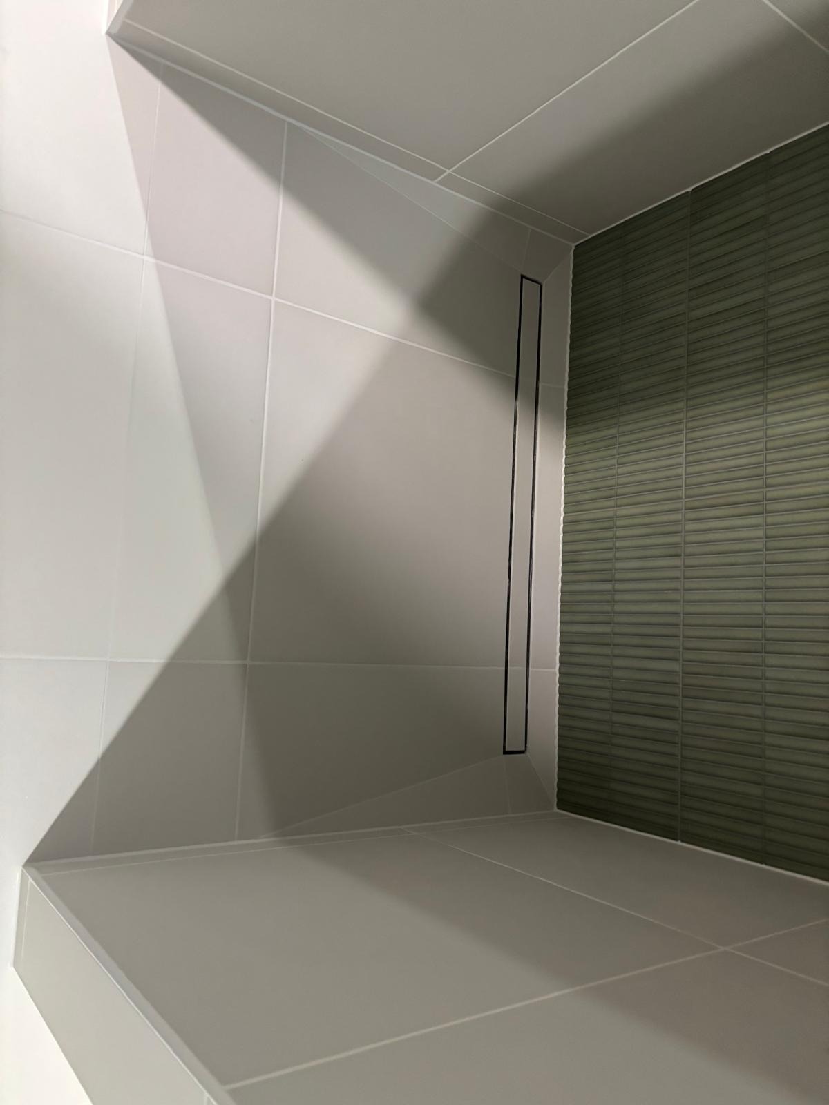 Nowoczesna kabina prysznicowa z odpływem liniowym i zieloną mozaiką na ścianie. Minimalistyczny design, jasne płytki, precyzyjne fugi.