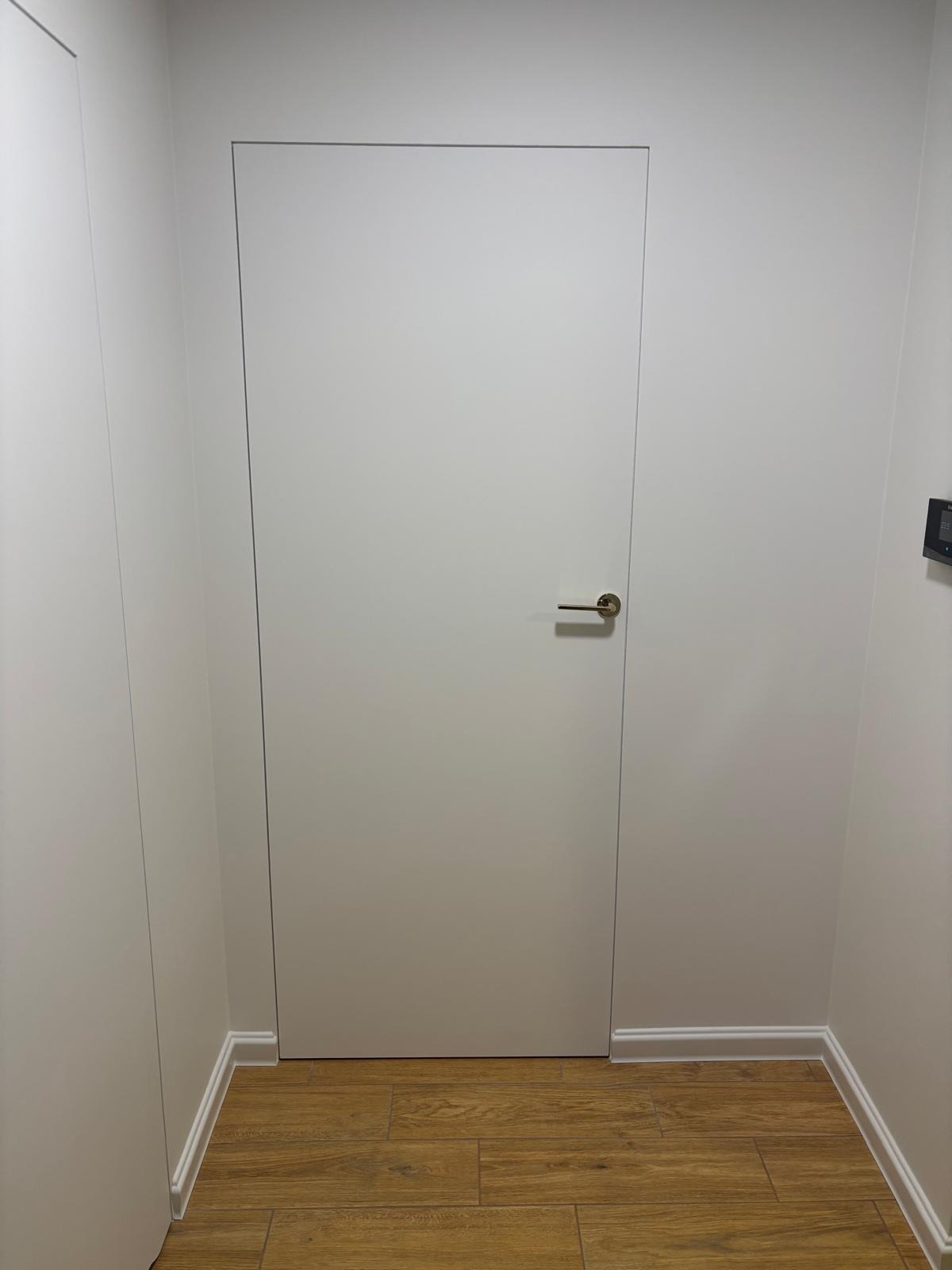 Minimalistyczne wnętrze: białe ściany, ukryte drzwi z dyskretną klamką i podłoga z paneli imitujących drewno. Nowoczesne wykończenie, subtelne detale.