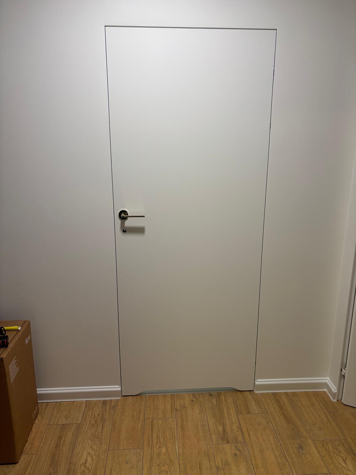Białe, minimalistyczne drzwi ukryte w ścianie, z widoczną srebrną klamką, na tle drewnianej podłogi i białych listew przypodłogowych. Częściowo widoczny karton z narzędziami.