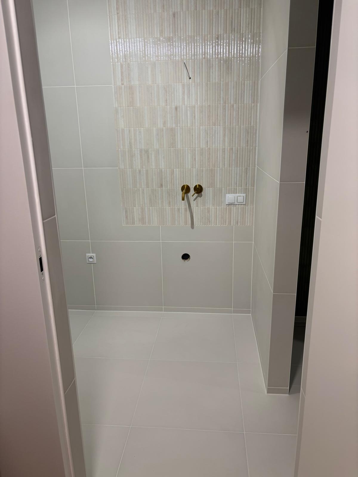 Wykończona łazienka z beżowymi płytkami imitującymi cegłę nad złotą armaturą. Widoczne gniazdko i otwór na rurę. Minimalistyczny design, jasne płytki podłogowe.