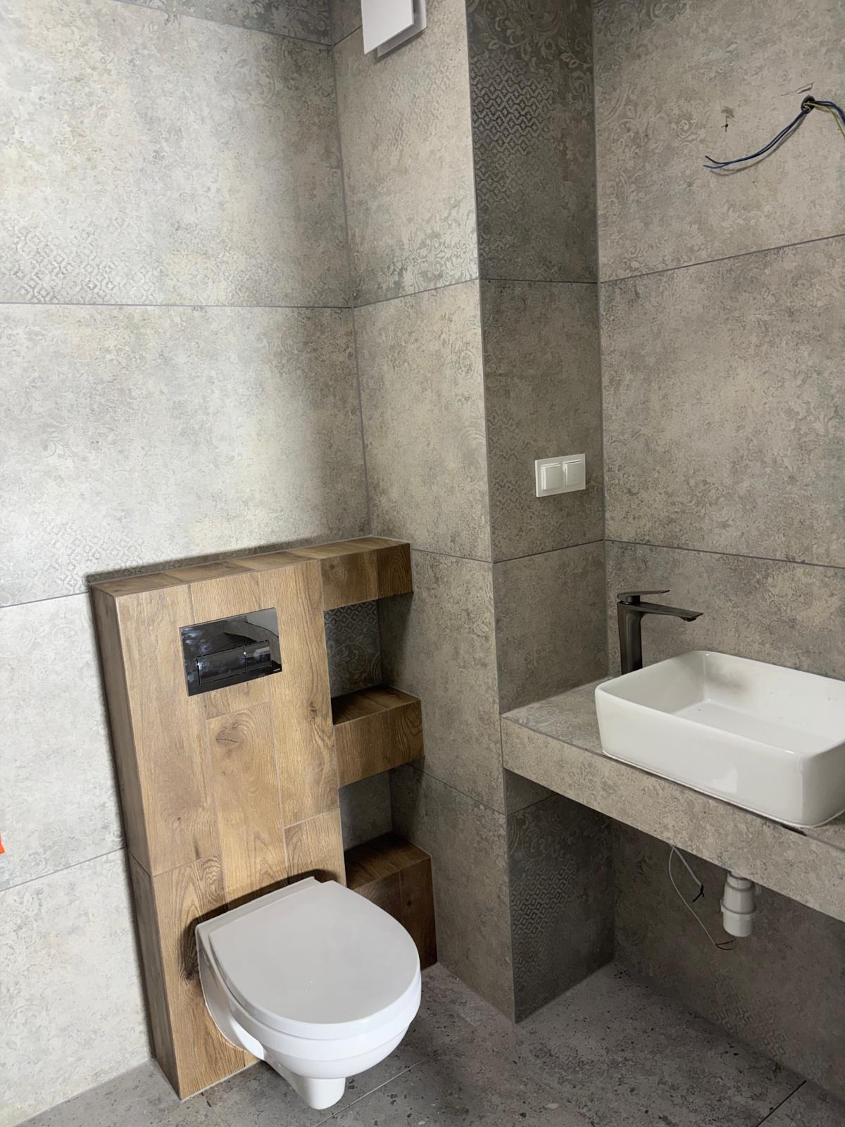 Nowoczesna łazienka z szarymi płytkami w ornamenty, drewnianą zabudową WC i białą umywalką nablatową z czarną armaturą. Widoczne kable elektryczne.