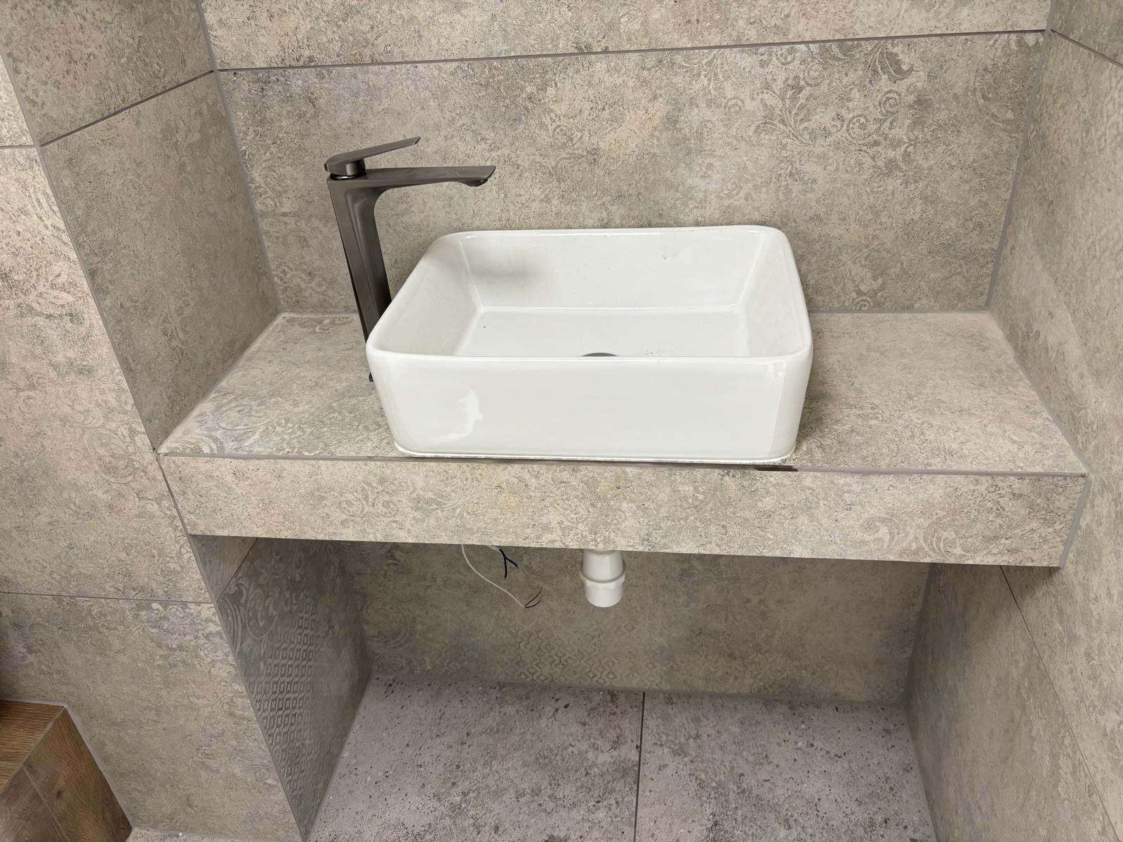 Kwadratowa, biała umywalka nablatowa z grafitową baterią, zamontowana na blacie z płytek w odcieniach szarości. Widoczne płytki na ścianie i pod blatem, minimalistyczny design.