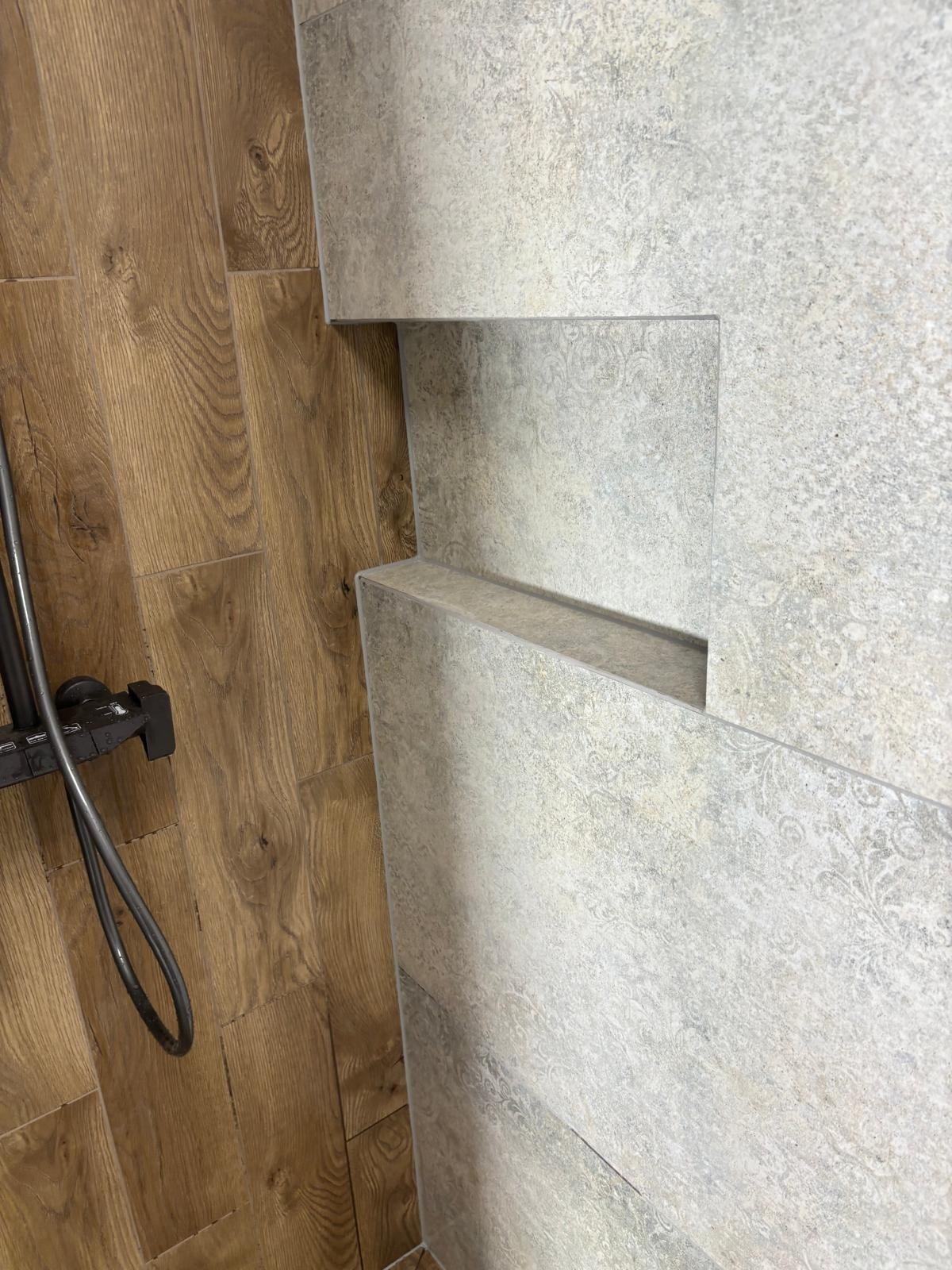 Wykończona łazienka z drewnopodobną podłogą i szarą, wzorzystą ścianą z płytek z wnęką. Widoczna bateria prysznicowa.