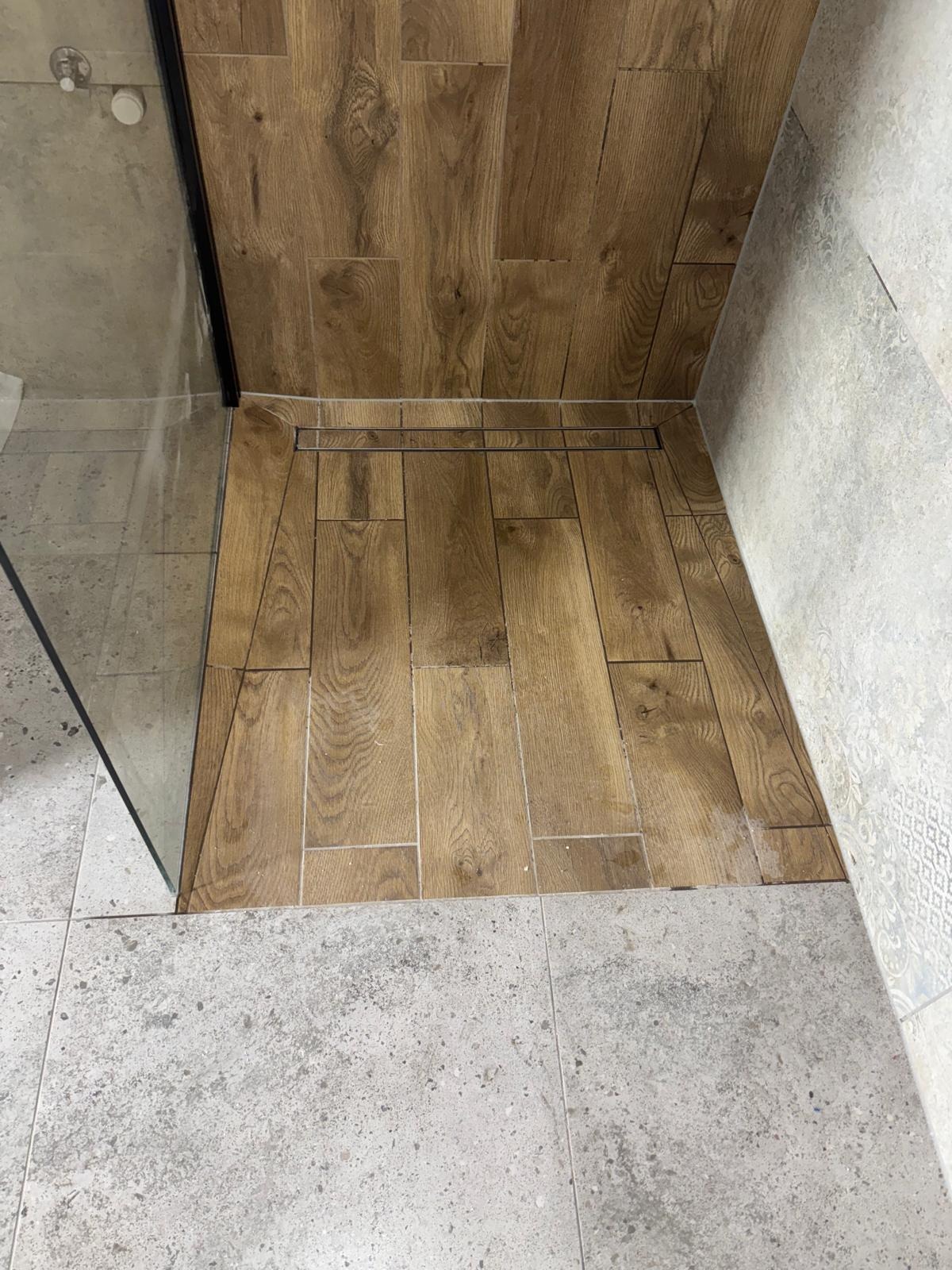 Wykończona strefa prysznicowa z odpływem liniowym, wyłożona płytkami imitującymi drewno, kontrastująca z szarą, wzorzystą okładziną ścienną i podłogową.