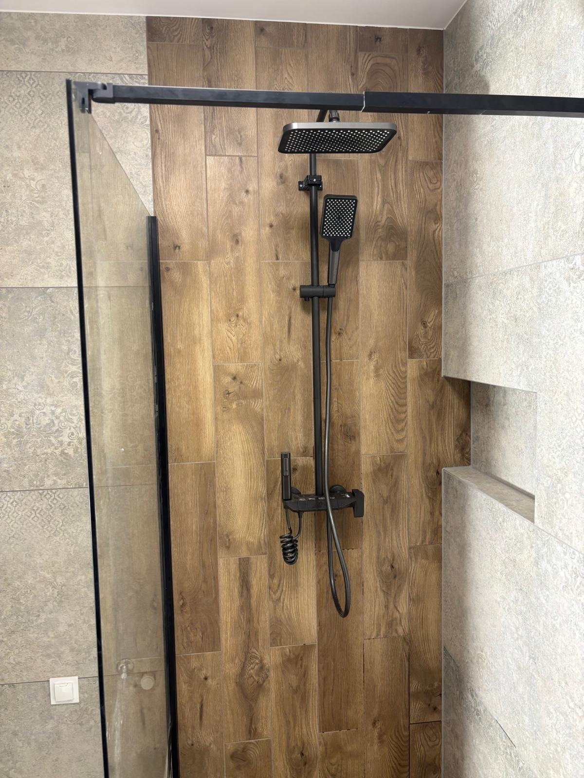 Kabina prysznicowa z czarną armaturą na tle drewnopodobnych płytek. Widoczny panel prysznicowy z deszczownicą i słuchawką. Nowoczesny design łazienki po remoncie.