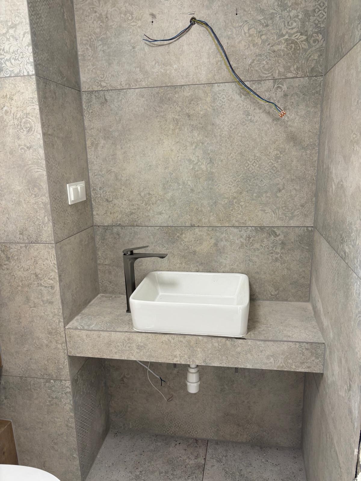 Wykończona łazienka z umywalką nablatową i baterią w kolorze czarnym, płytki w odcieniach szarości z delikatnym wzorem. Widoczne przewody elektryczne.