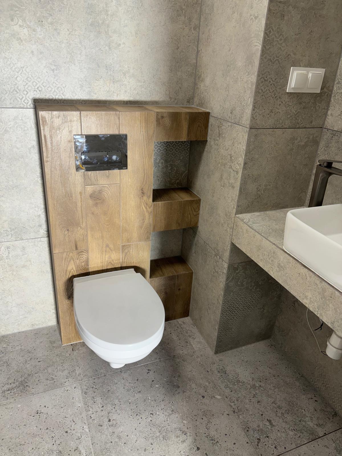 Nowoczesna toaleta podwieszana z drewnianą obudową i półkami w łazience wykończonej szarą, wzorzystą płytką. Umywalka nablatowa z szarą szafką.