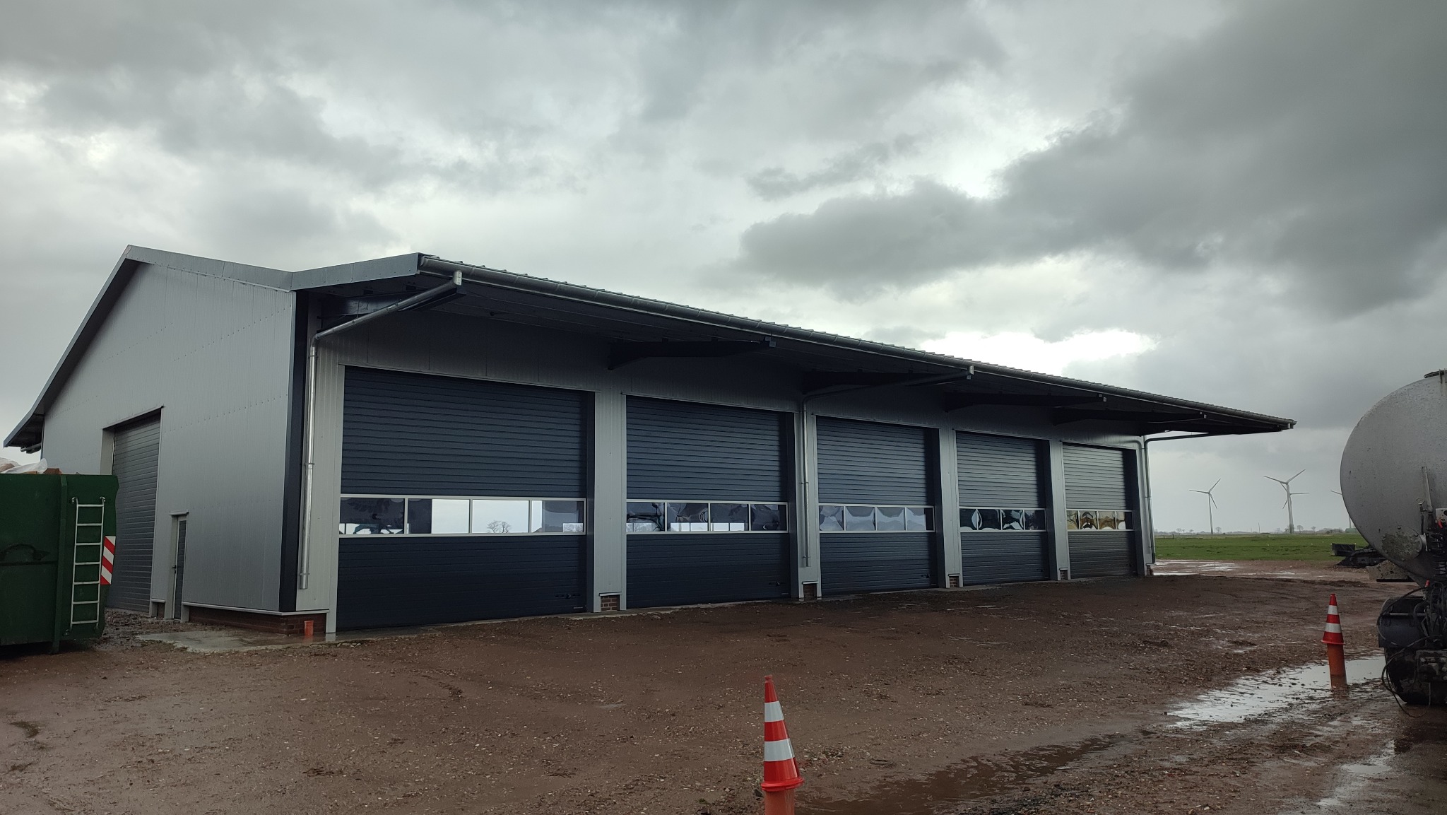 Nowoczesna hala z szarej blachy z trzema segmentowymi bramami garażowymi w kolorze antracytowym, widoczne turbiny wiatrowe w tle, pochmurne niebo.