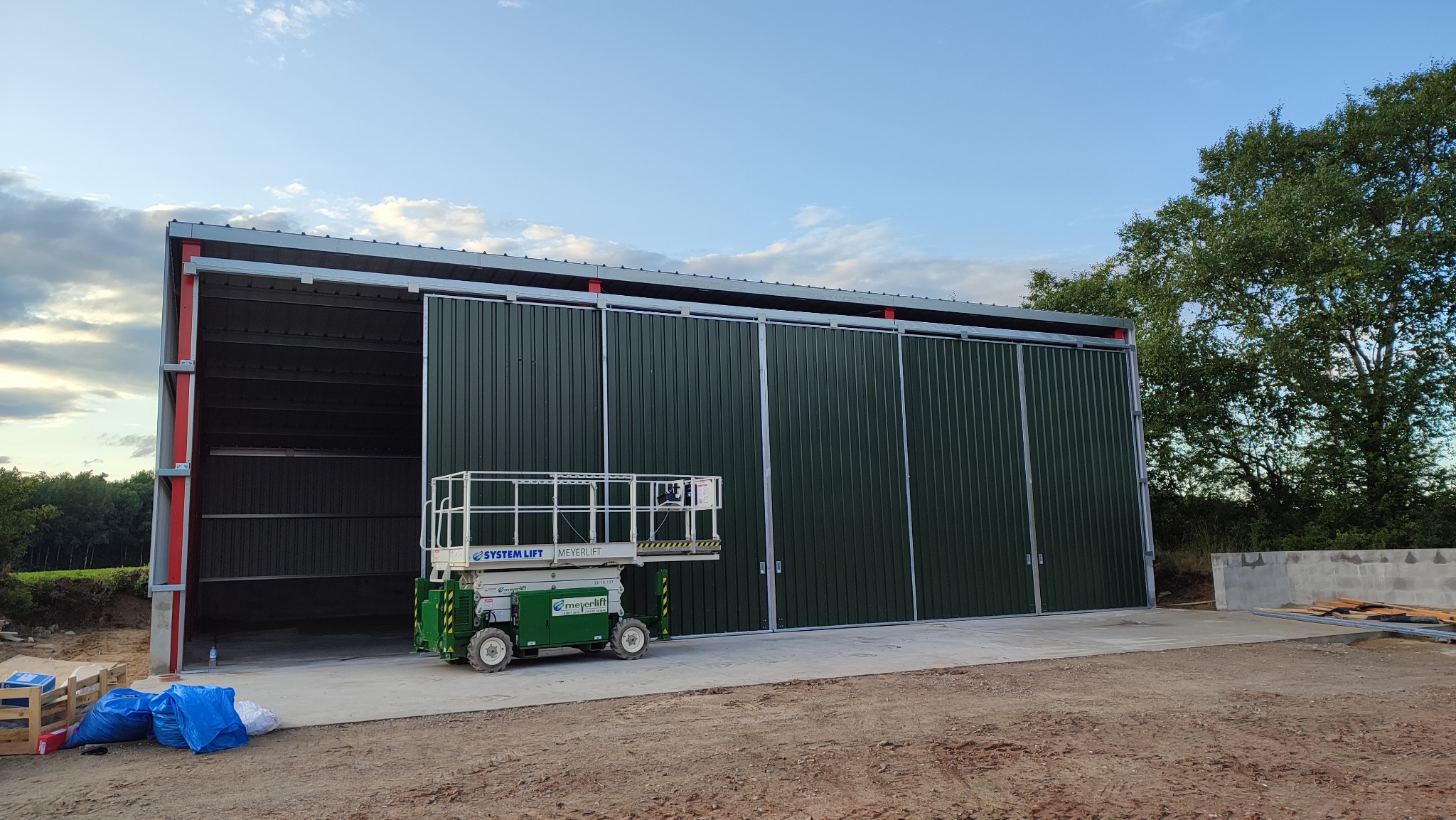 Zakończona budowa hali z zielonej blachy trapezowej z przesuwnymi drzwiami, widoczny podnośnik nożycowy System Lift i materiały budowlane na zewnątrz.