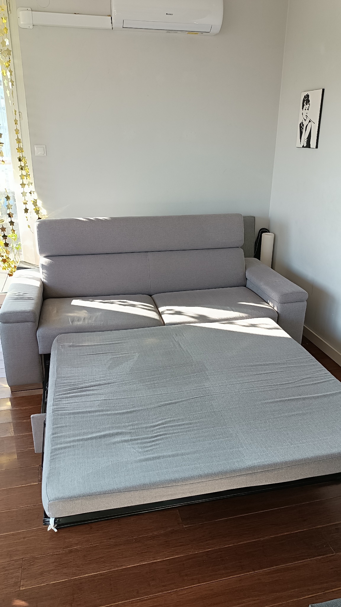 Rozłożona szara kanapa z wysuwanym siedziskiem na drewnianej podłodze, widok z przodu w jasnym pokoju z dekoracją okienną i obrazem na ścianie.