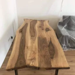 Stół z blatem z litego drewna o nieregularnych krawędziach i metalowych nogach, widoczne słoje drewna, w tle ściana i zakryta folią kanapa.