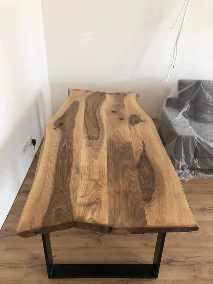 Stół z blatem z litego drewna o nieregularnych krawędziach i metalowych nogach, widoczne słoje drewna, w tle ściana i zakryta folią kanapa.