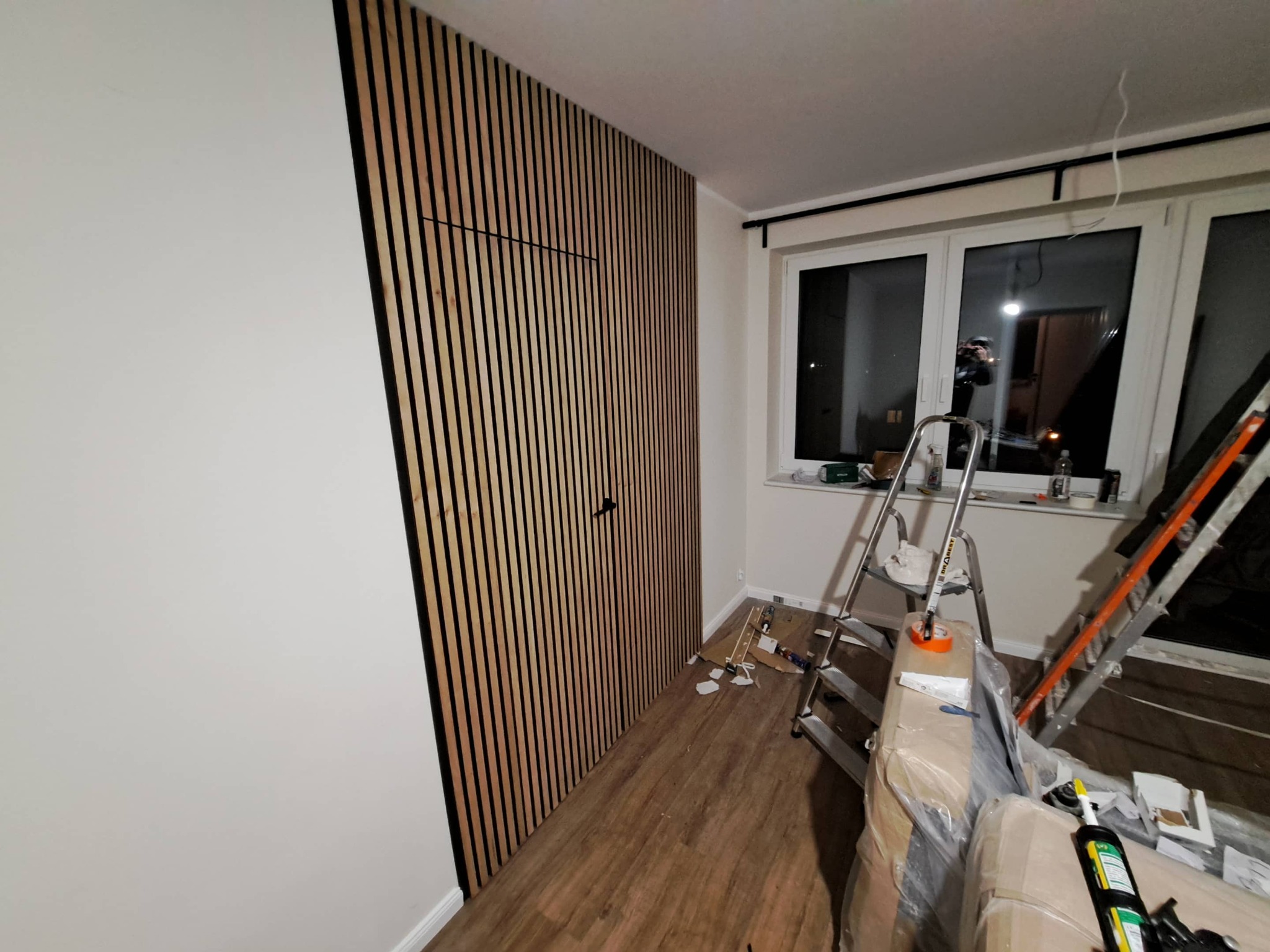 Ściana z drewnianych lameli z ukrytymi drzwiami, widoczne narzędzia i drabina, podłoga drewniana. Część ściany pomalowana na biało. Nowoczesny design wnętrza.