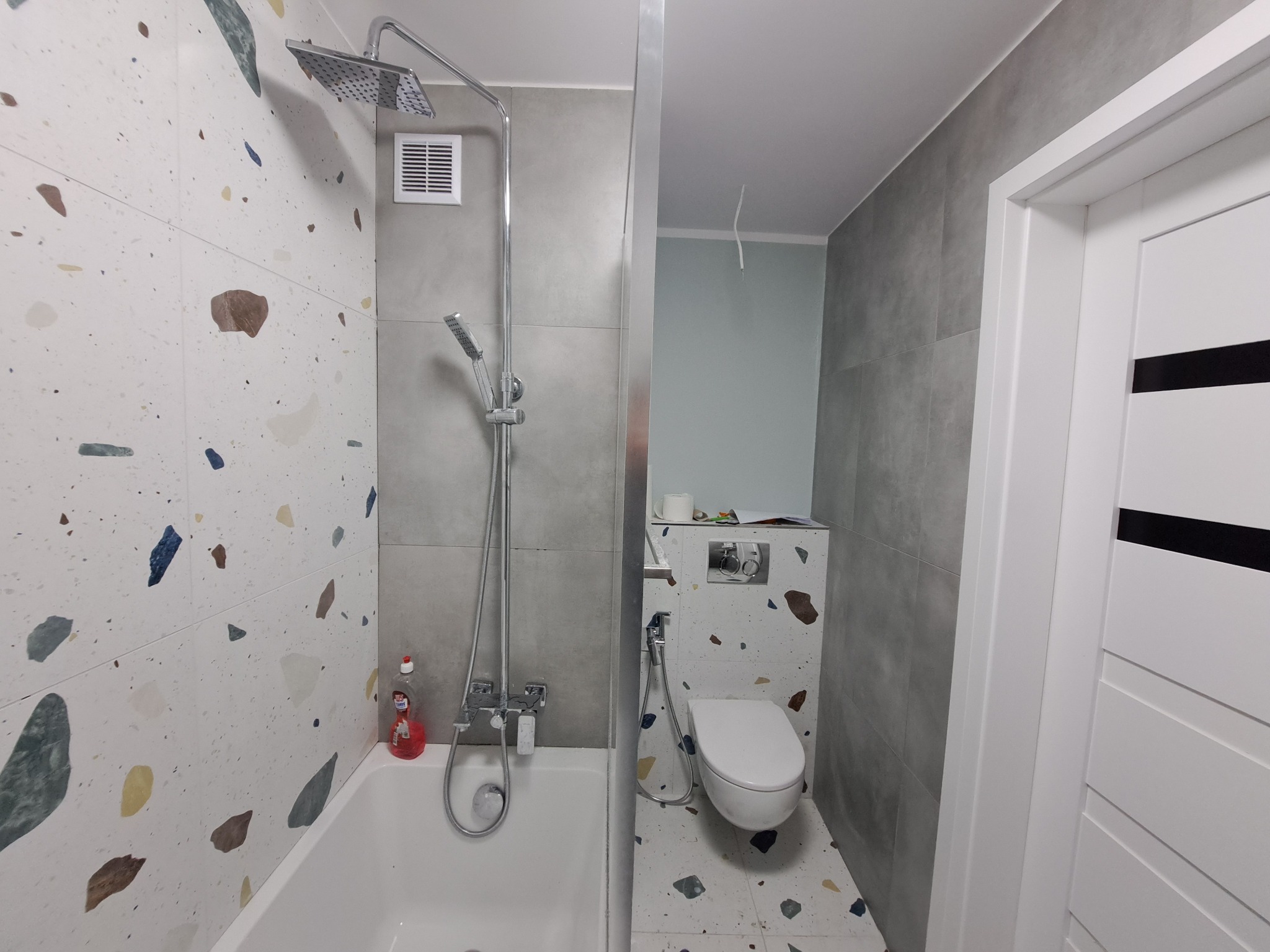 Wykończona łazienka z wanną, prysznicem i toaletą. Ściany wyłożone płytkami w dwóch odcieniach szarości i wzorzystymi płytkami imitującymi lastryko. Białe drzwi z czarnym akcentem.