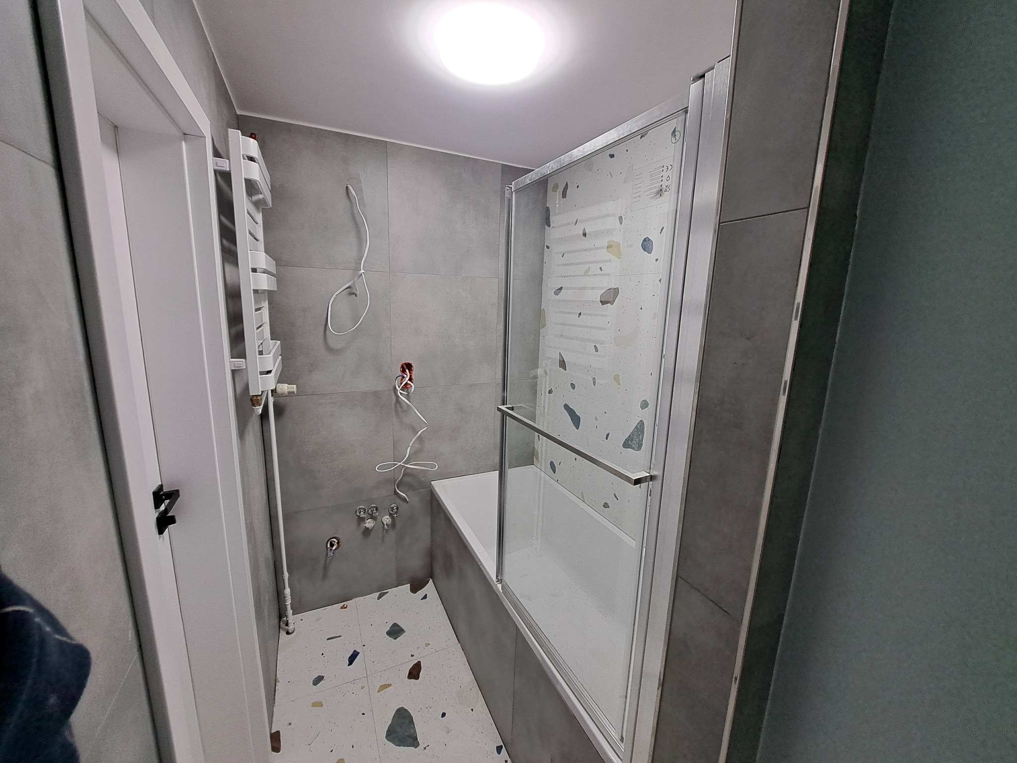 Nowoczesna łazienka w szarościach z wanną za szklanym parawanem, mozaikowa podłoga, grzejnik drabinkowy i przygotowane instalacje pod baterie.