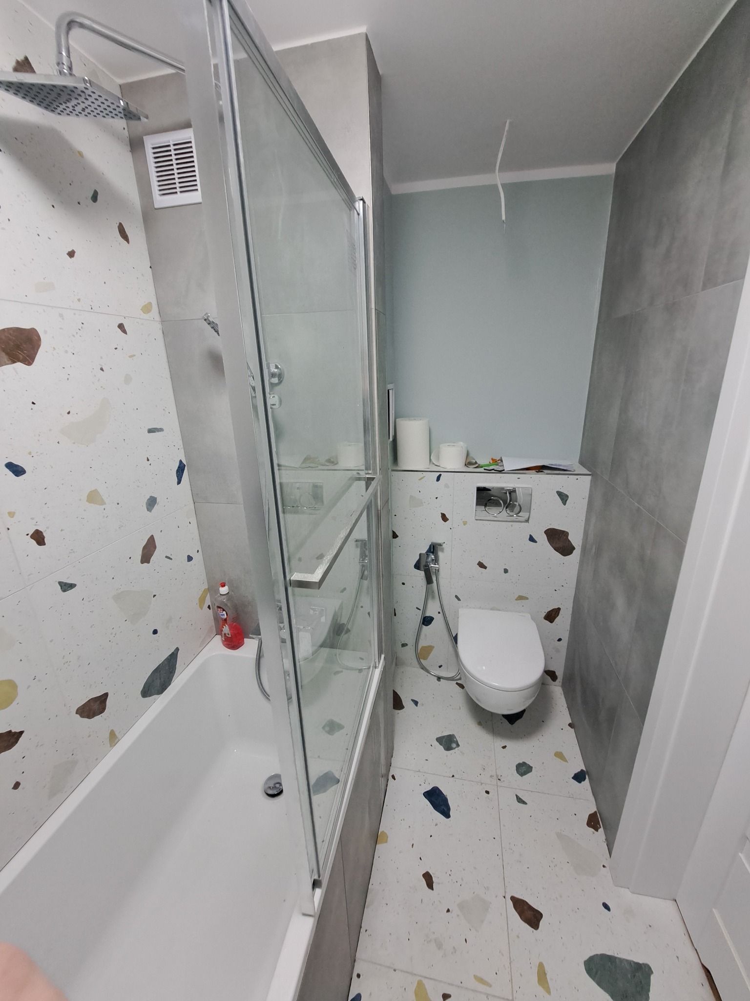 Wykończona łazienka z wanną, kabiną prysznicową i toaletą. Na ścianach i podłodze płytki w stylu lastryko. Ściana nad toaletą pomalowana na jasny, pastelowy kolor.