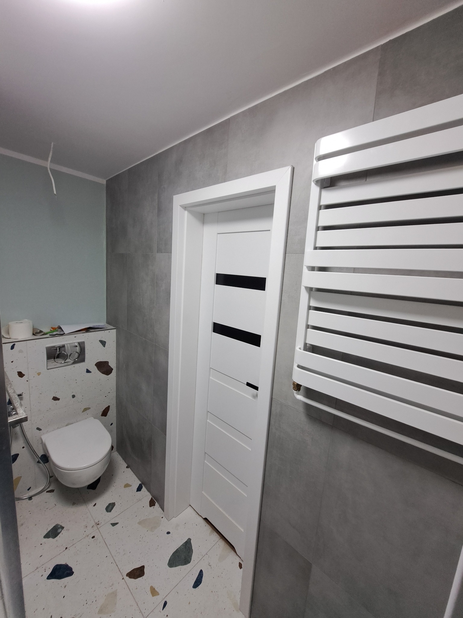 Nowoczesna łazienka po remoncie: szare płytki ścienne, biała armatura, grzejnik drabinkowy i drzwi z czarnymi akcentami. Podłoga w stylu lastryko.