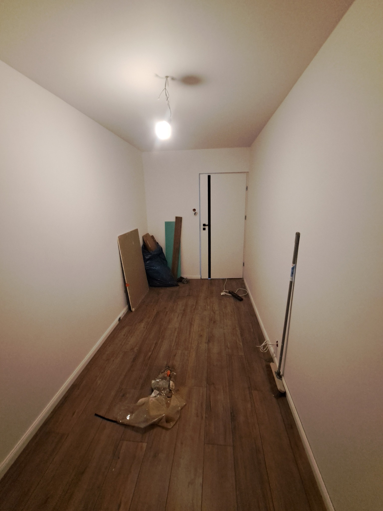 Korytarz po remoncie z panelami podłogowymi imitującymi drewno, białe ściany, białe drzwi z czarnym akcentem. Widoczne ślady po remoncie: worki, płyty, narzędzia.