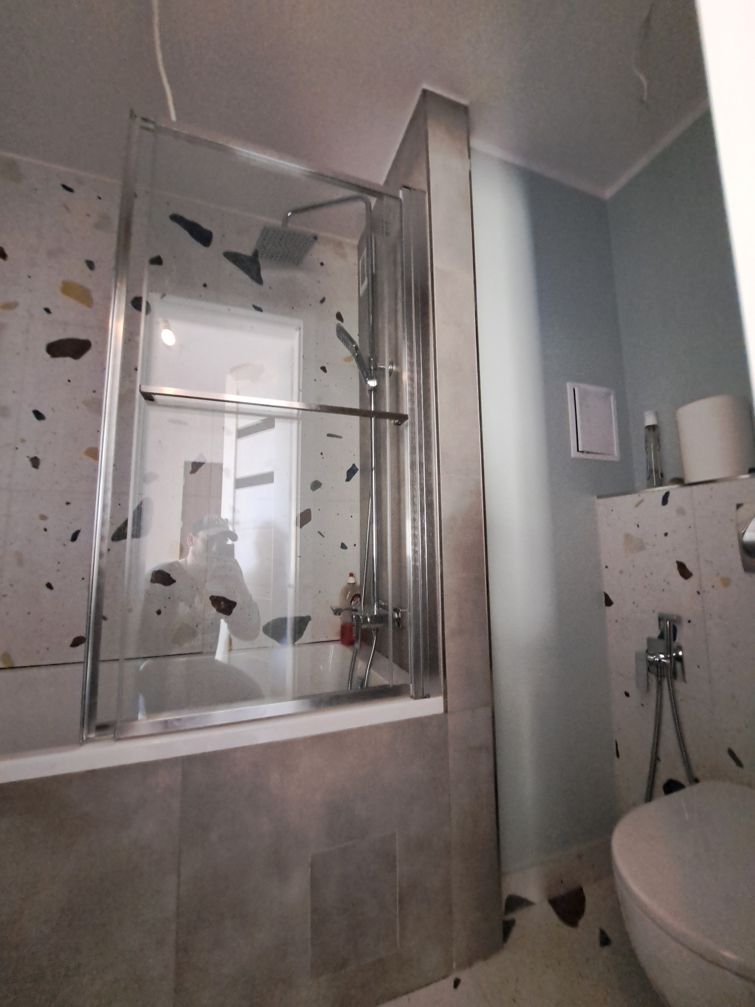 Nowoczesna kabina prysznicowa z transparentnego szkła, zamontowana nad wanną w łazience wykończonej płytkami w stylu lastryko. Widoczna armatura i toaleta.