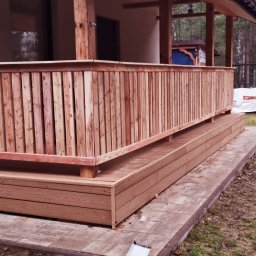 Łukasz - Drewniana balustrada z pionowych desek, zamontowana na tarasie przy domu. Widoczne elementy konstrukcyjne i wykończeniowe. Tło stanowią drzewa i fragment budynku.