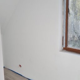 Łukasz - Wnętrze po malowaniu: białe ściany, drewnopodobna podłoga zabezpieczona folią malarską, okno w drewnianej ramie. Widoczny fragment ściany ze skośnym sufitem.