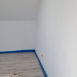 Łukasz - Jasne panele podłogowe w pokoju z białymi ścianami, zabezpieczone niebieską folią malarską przy listwach. W rogu leżą elementy wykończeniowe.