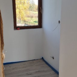 Łukasz - Pokój po remoncie: szare panele podłogowe, białe ściany z brązową ramą okienną. Widoczne zabezpieczenia taśmą malarską przy podłodze i wystające kable elektryczne.