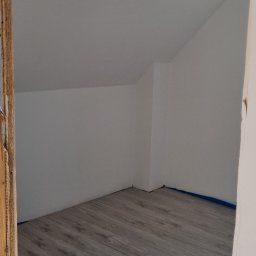 Łukasz - Poddasze z oknem dachowym i panelami podłogowymi w odcieniach szarości. Ściany pomalowane na biało, zabezpieczone niebieską taśmą malarską przy podłodze.