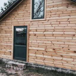 Łukasz - Ściana budynku obita jasnym drewnem z widocznymi oknami i drzwiami w ciemnej ramie. Fragment ściany z białych bloczków. Ułożone schodki z kostki.