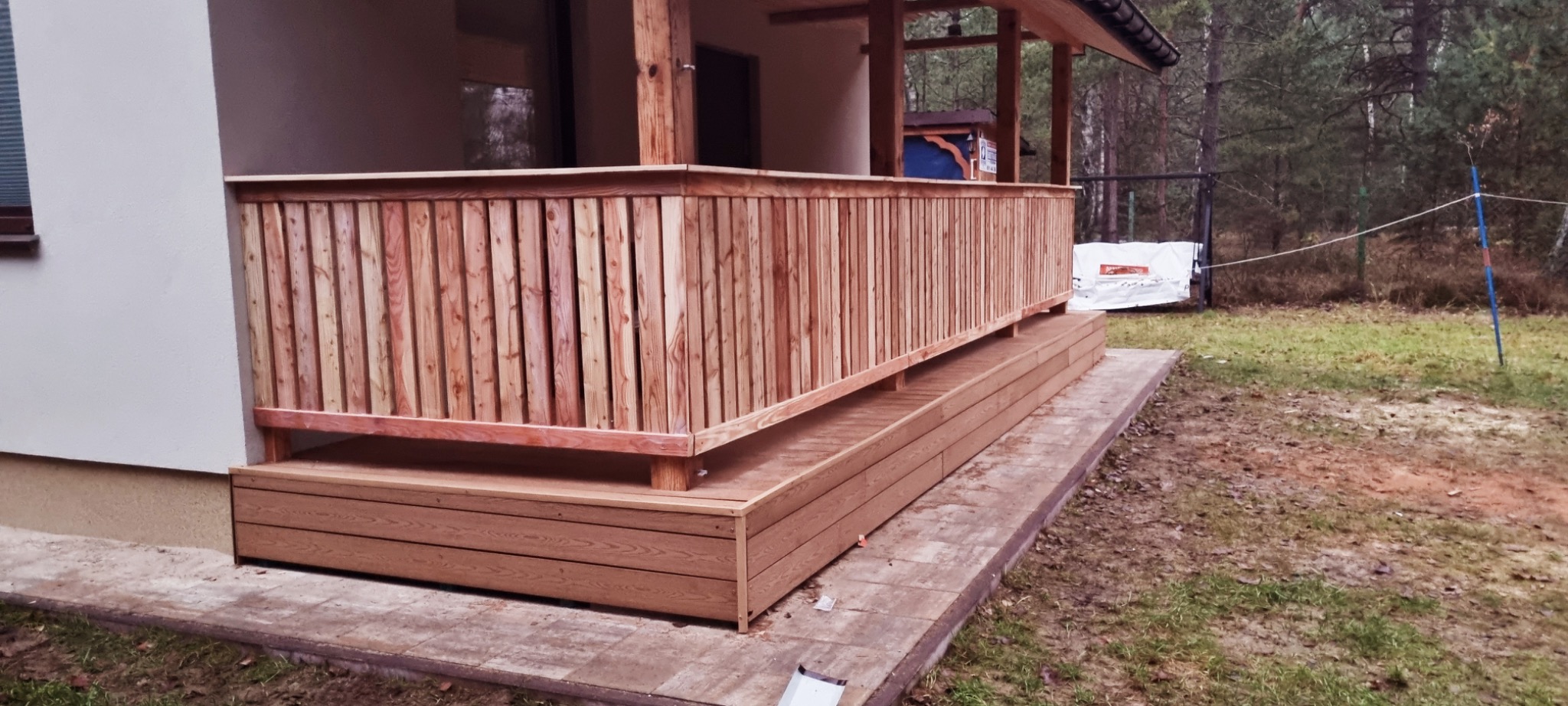 Drewniana balustrada z pionowych desek, zamontowana na tarasie przy domu. Widoczne elementy konstrukcyjne i wykończeniowe. Tło stanowią drzewa i fragment budynku.