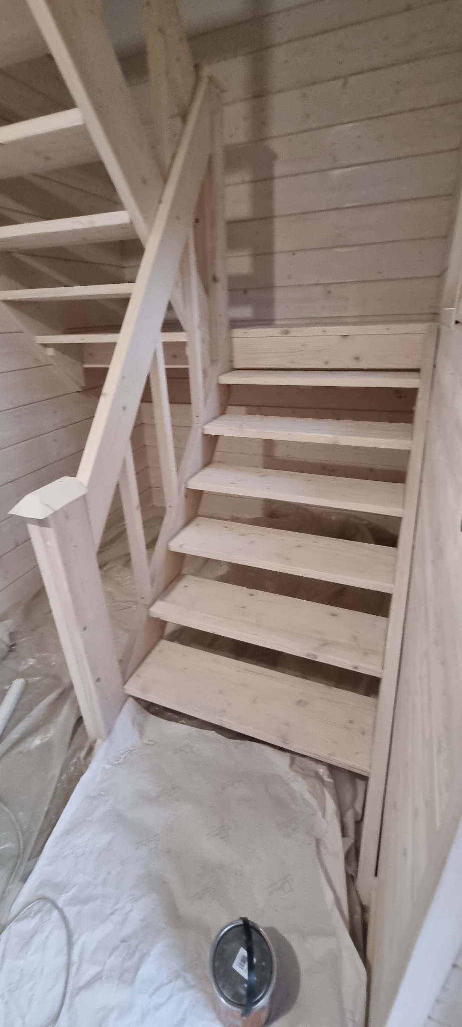 Jasne, drewniane schody z balustradą w domu o ścianach z boazerii. Widoczna puszka z farbą i folia ochronna na podłodze.