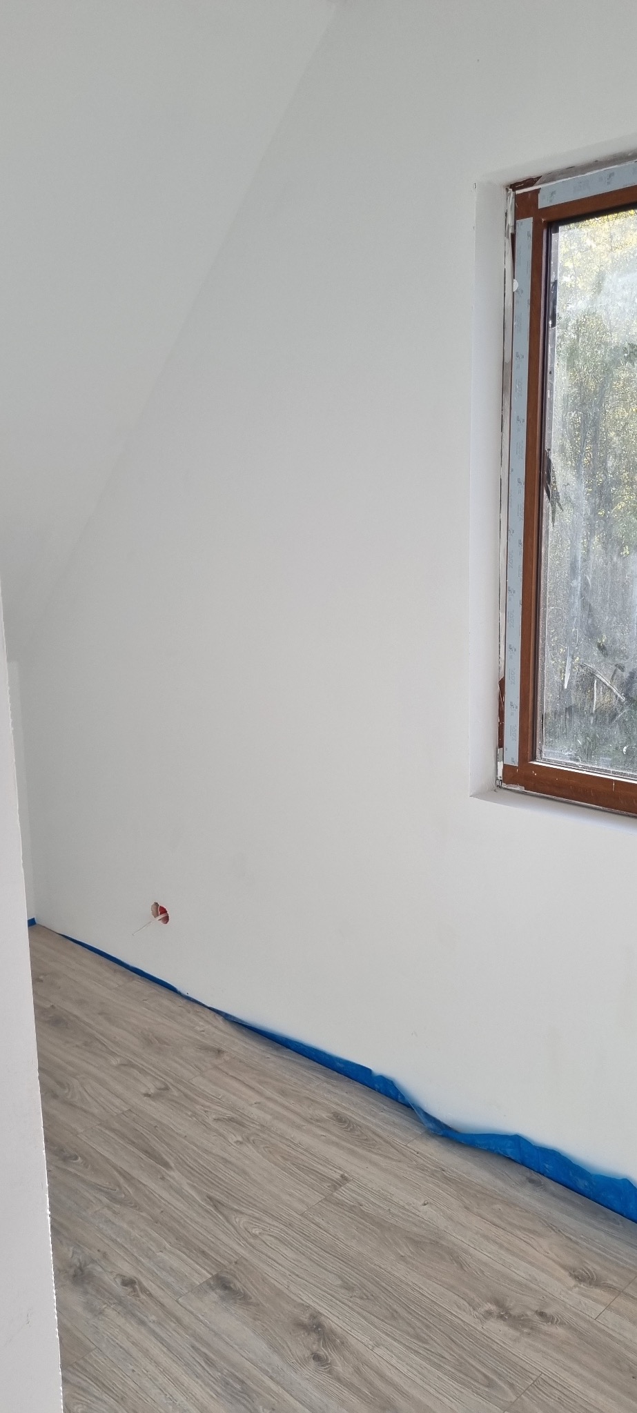 Wnętrze po malowaniu: białe ściany, drewnopodobna podłoga zabezpieczona folią malarską, okno w drewnianej ramie. Widoczny fragment ściany ze skośnym sufitem.