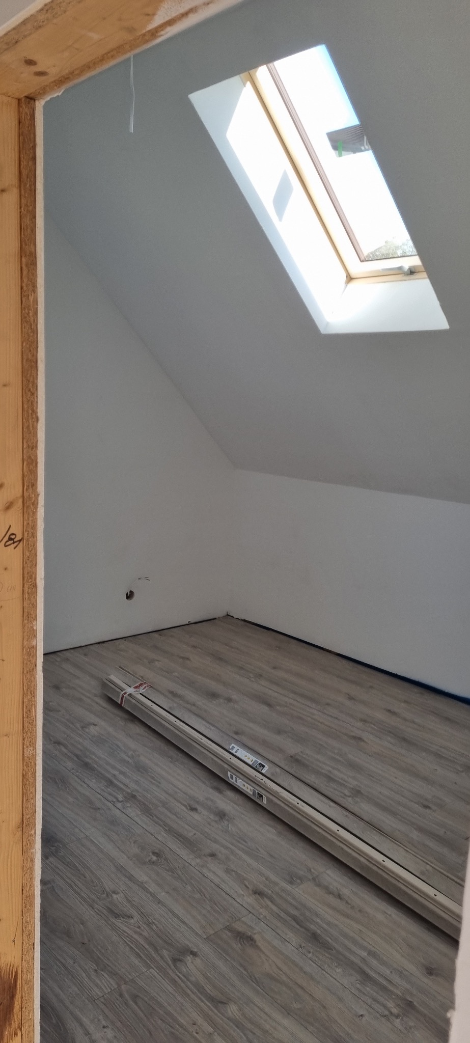 Poddasze z oknem dachowym i panelami podłogowymi przed montażem. Widoczna surowa rama drzwi i świeżo pomalowane ściany. Podłoga w odcieniach szarości.