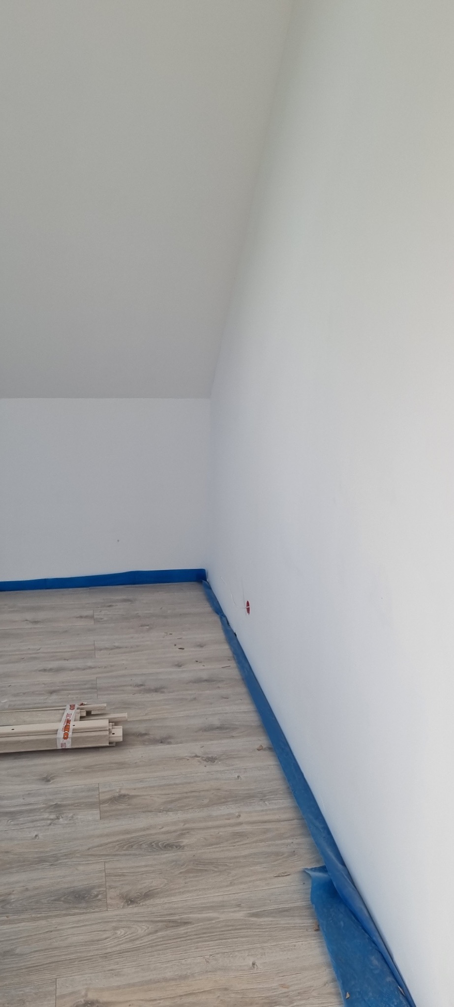 Jasne panele podłogowe w pokoju z białymi ścianami, zabezpieczone niebieską folią malarską przy listwach. W rogu leżą elementy wykończeniowe.