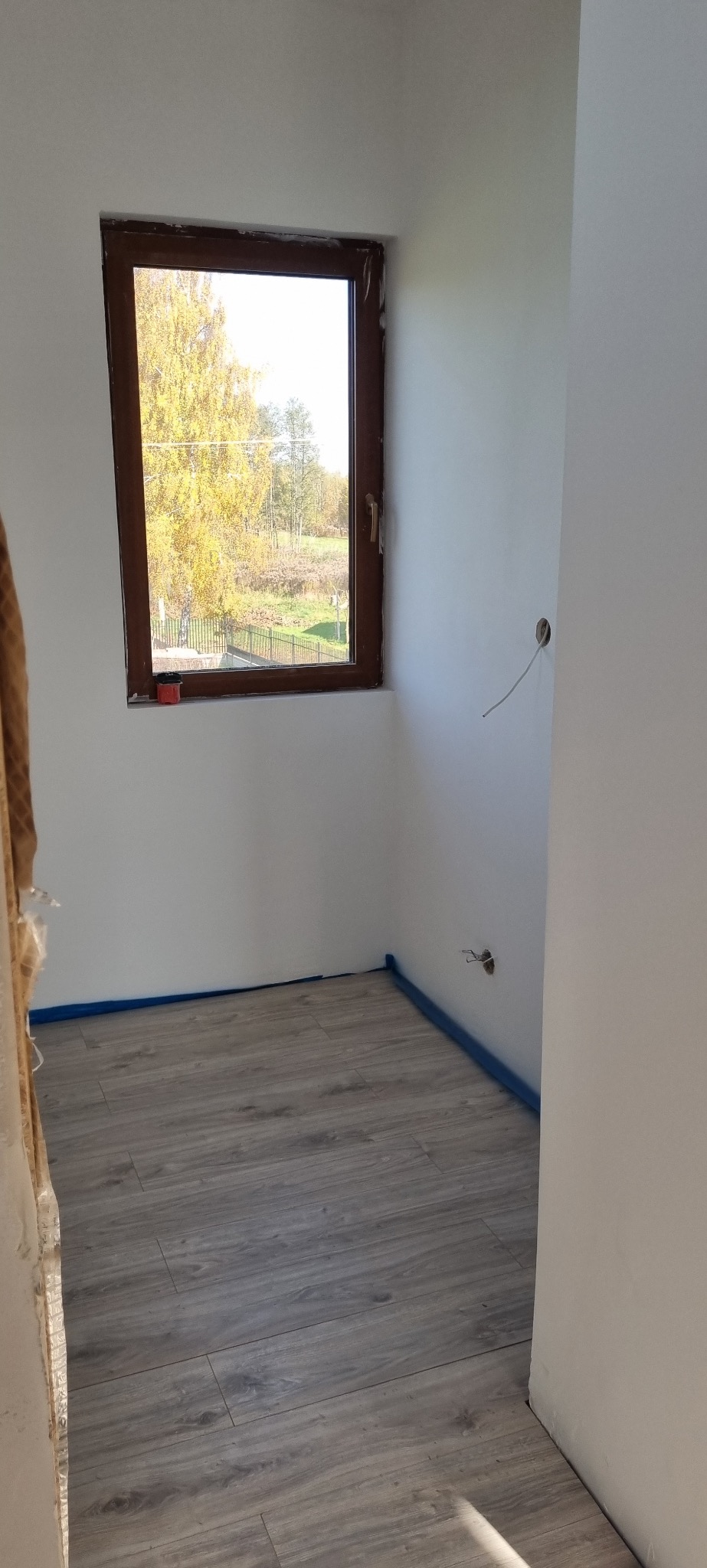 Pokój po remoncie: szare panele podłogowe, białe ściany z brązową ramą okienną. Widoczne zabezpieczenia taśmą malarską przy podłodze i wystające kable elektryczne.