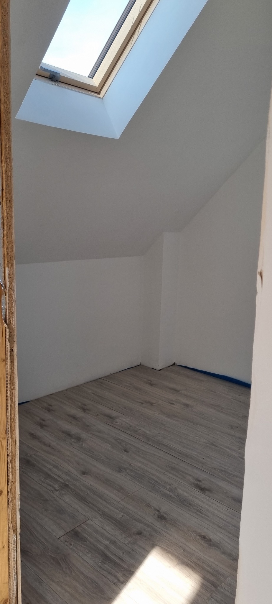 Poddasze z oknem dachowym i panelami podłogowymi w odcieniach szarości. Ściany pomalowane na biało, zabezpieczone niebieską taśmą malarską przy podłodze.