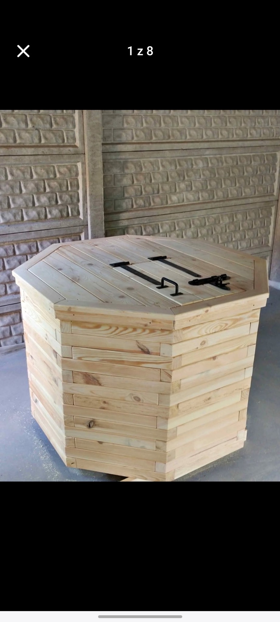 Drewniana studnia ogrodowa o ośmiokątnym kształcie, wykonana z jasnego drewna, z czarnymi metalowymi uchwytami, na tle betonowego ogrodzenia.