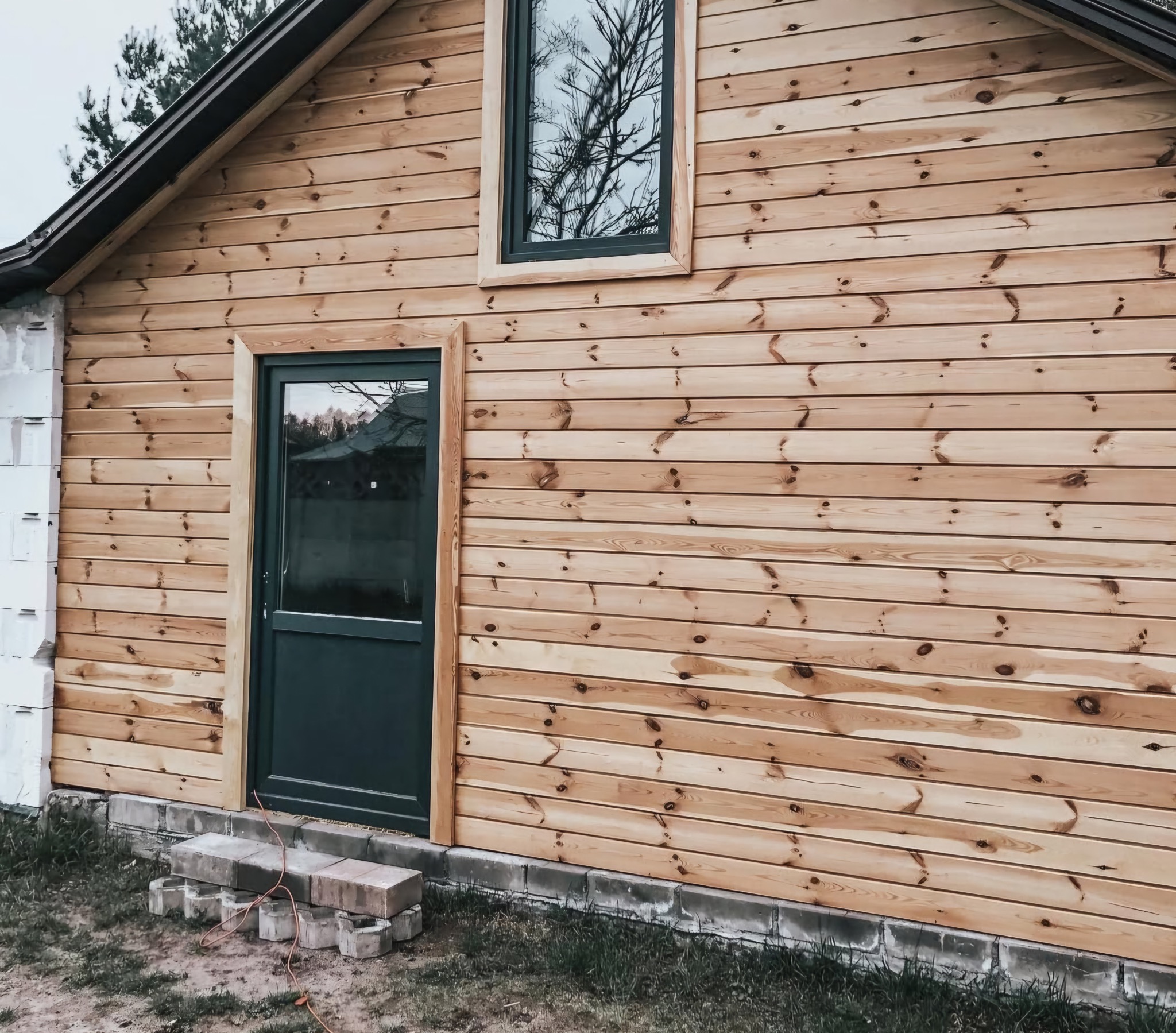 Ściana budynku obita jasnym drewnem z widocznymi oknami i drzwiami w ciemnej ramie. Fragment ściany z białych bloczków. Ułożone schodki z kostki.