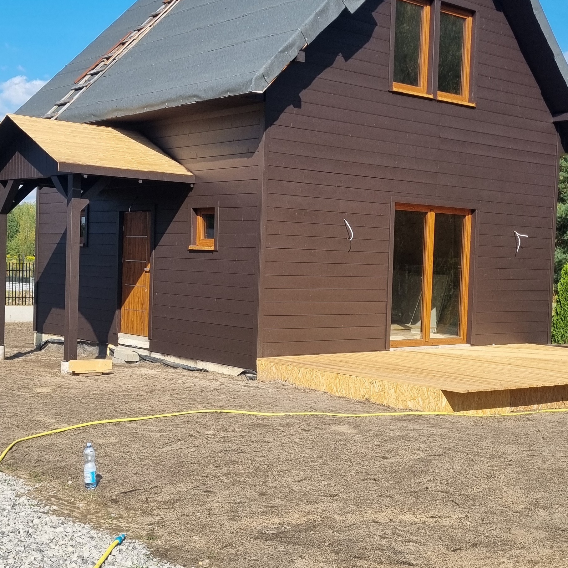Dom z ciemną elewacją i jasnym drewnianym tarasem. Widoczny fragment dachu z drabinką. Na pierwszym planie butelka wody i żółty wąż ogrodowy.