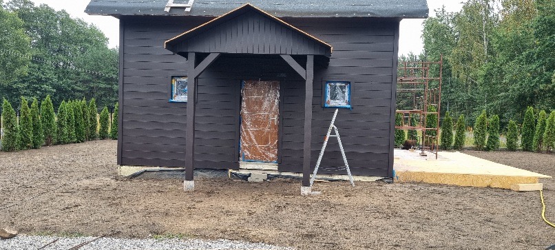 Ciemna altana z podestem w trakcie budowy, okna zabezpieczone folią, drabina oparta o ścianę, w tle drzewa i tuje. Widoczny etap prac wykończeniowych.