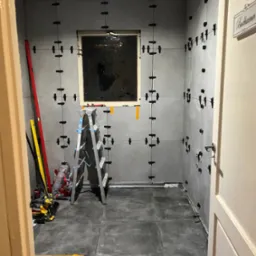 Szare płytki ścienne z czarnymi krzyżykami dystansowymi w trakcie układania w łazience, drabina aluminiowa, poziomice, narzędzia i worki z zaprawą w tle, widoczny fragment drzwi z tabliczką...