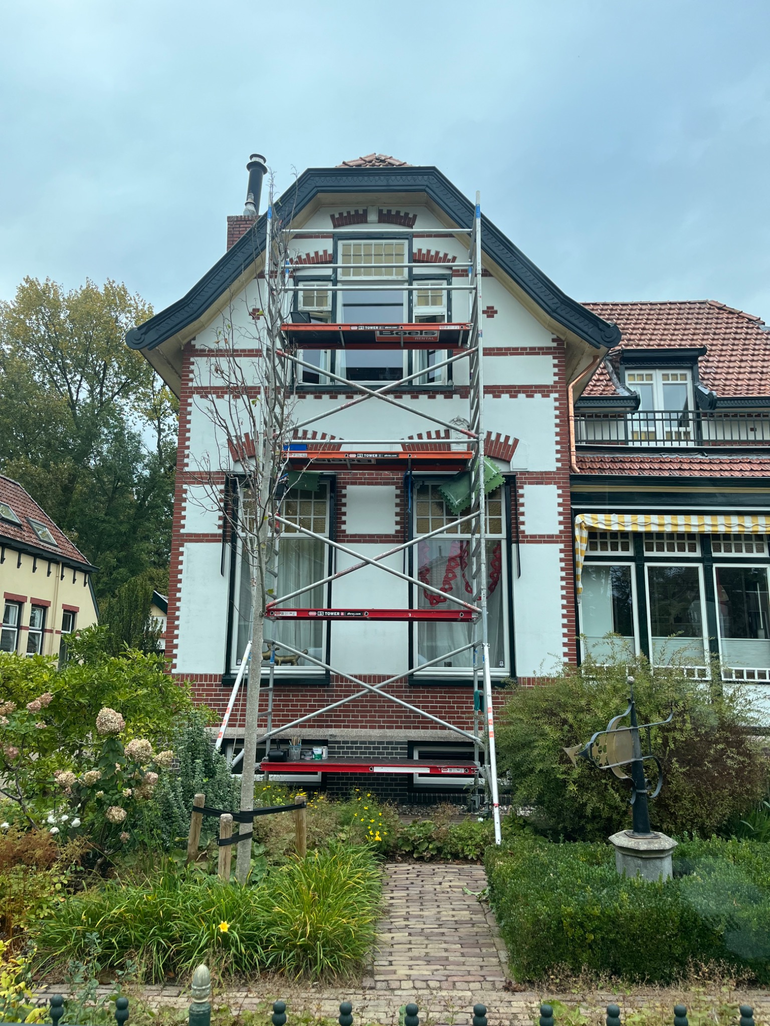 Dwupiętrowy dom z białymi ścianami i czerwoną cegłą, podczas prac remontowych, z rusztowaniem ustawionym przed fasadą, widoczne narzędzia na rusztowaniu.