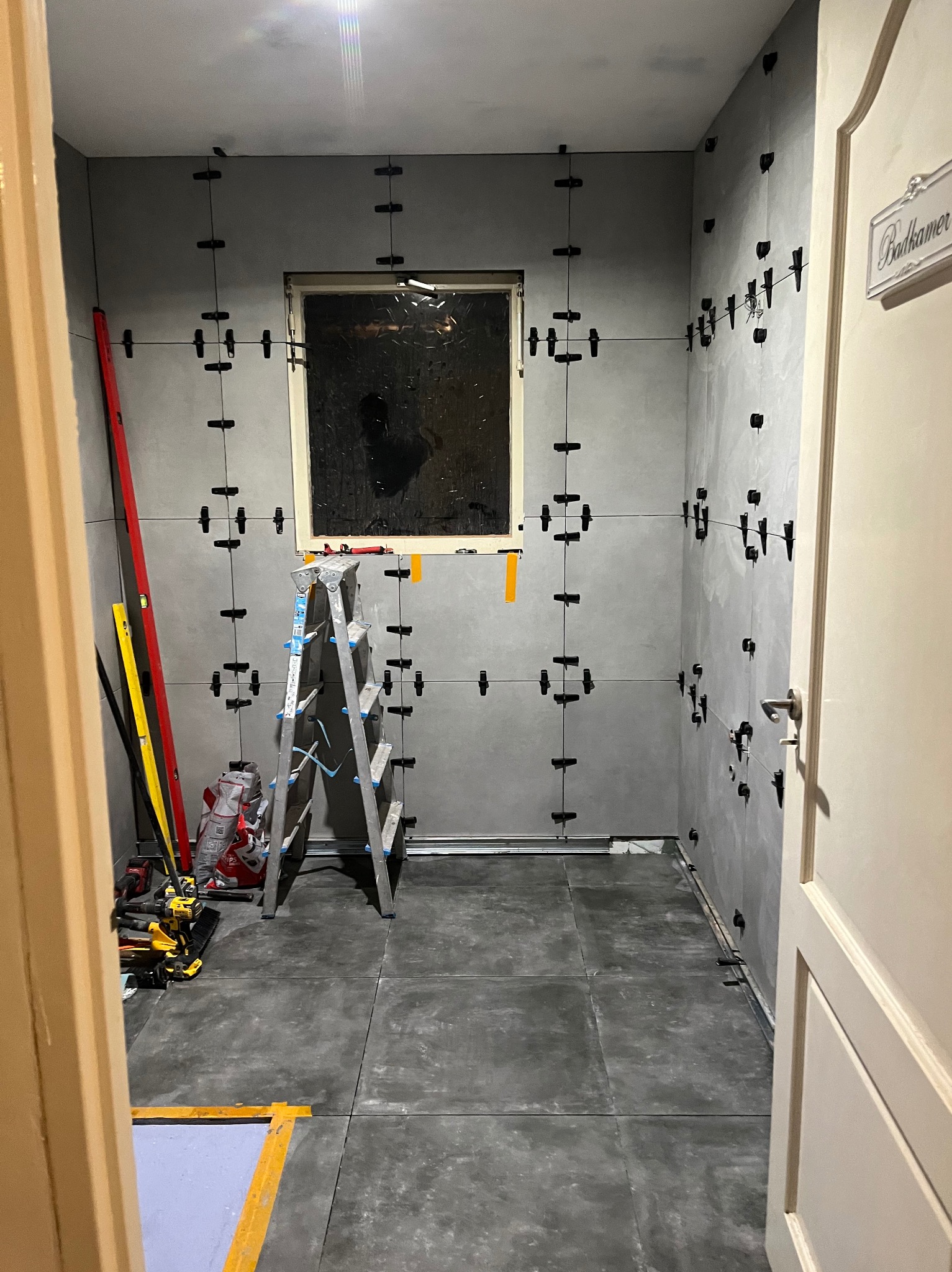 Szare płytki ścienne z czarnymi krzyżykami dystansowymi w trakcie układania w łazience, drabina aluminiowa, poziomice, narzędzia i worki z zaprawą w tle, widoczny fragment drzwi z tabliczką...