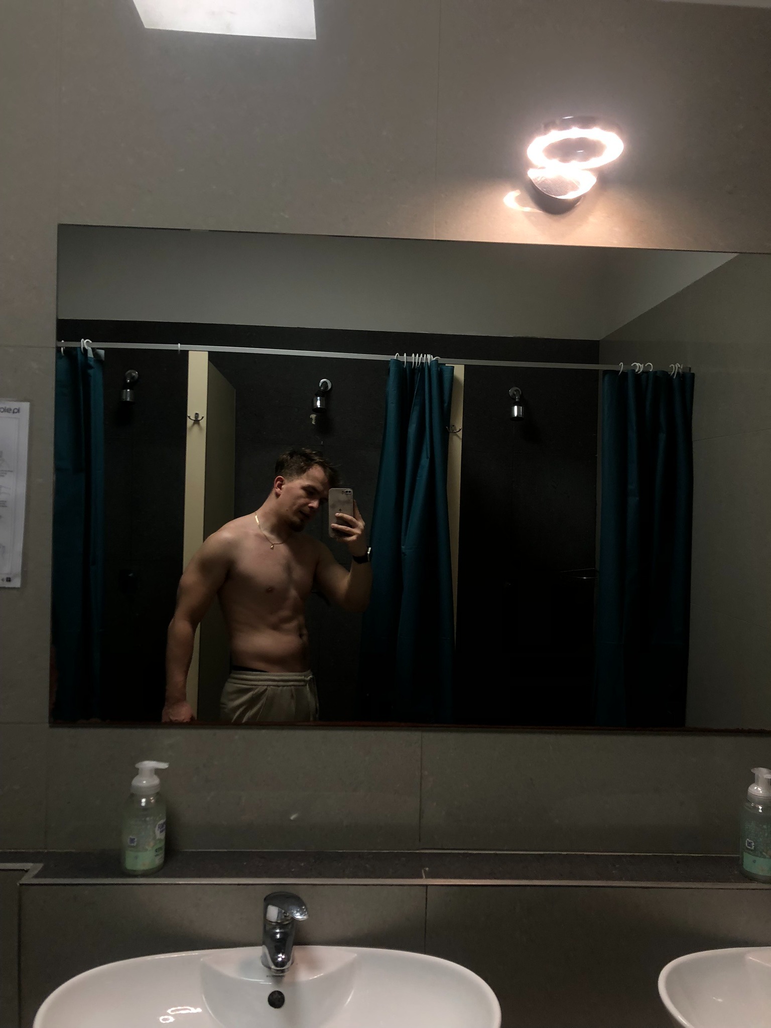 Mężczyzna bez koszulki robi selfie w lustrze w szatni siłowni, widoczny zarys mięśni brzucha, w tle kabiny prysznicowe z zasłonami.