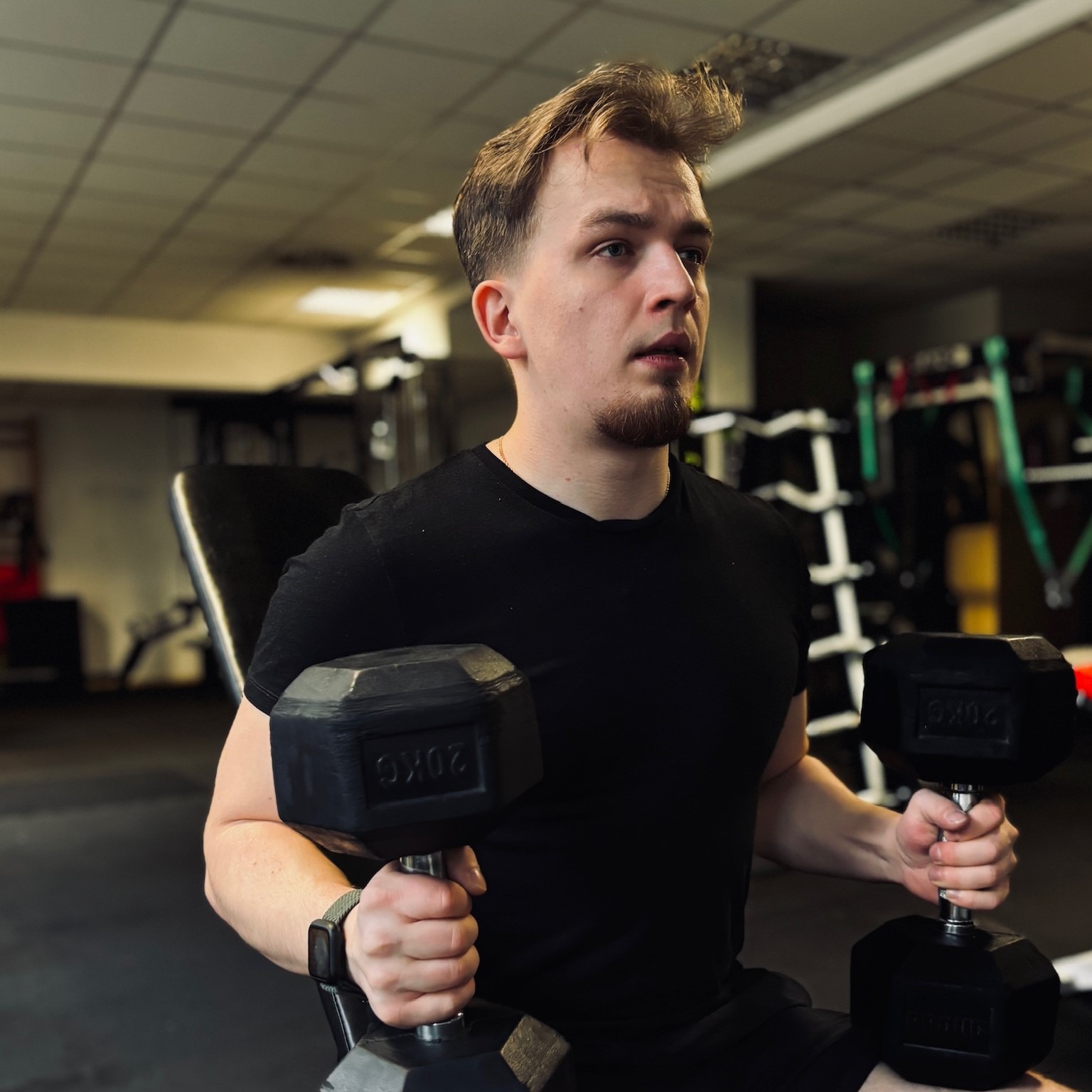 Młody mężczyzna w czarnej koszulce siedzi na ławce w siłowni, trzymając w rękach dwie 20-kilogramowe hantle, z wyrazem skupienia na twarzy.