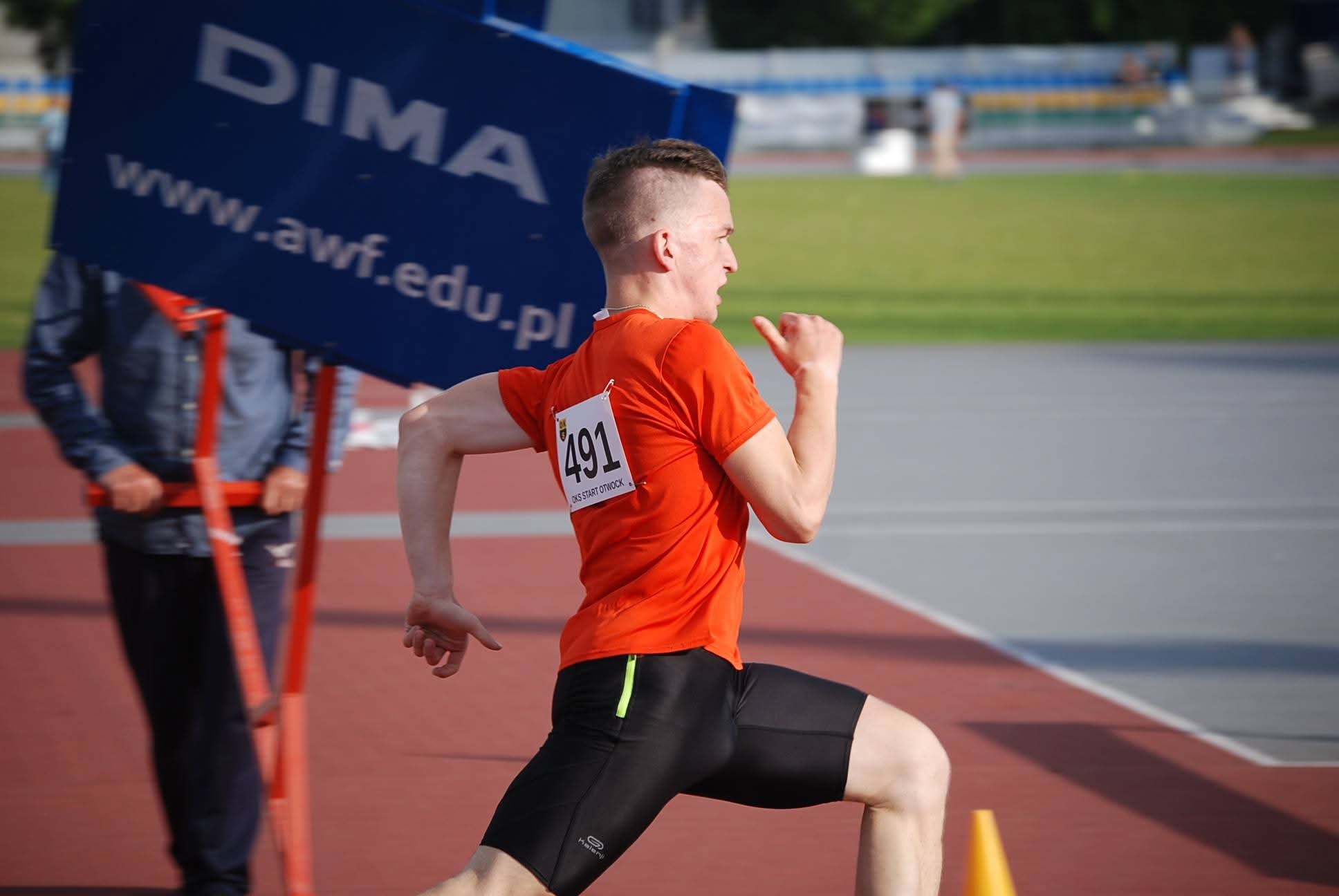 Biegacz w pomarańczowej koszulce z numerem 491, w trakcie sprintu na bieżni, widok z boku. W tle tablica informacyjna i zielone boisko.
