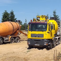 Wytwórnia Materiałów Budowlanych Zdzisława Szczepańska - Dwa samochody betonomieszarki, pomarańczowy Mercedes i żółty MAN, stoją na placu budowy obok wykopu z przygotowanym zbrojeniem fundamentów, gdzie robotnicy wylewają beton.