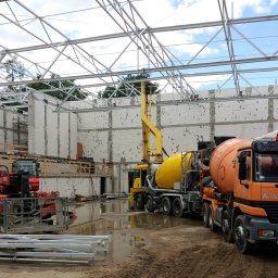 Wytwórnia Materiałów Budowlanych Zdzisława Szczepańska - Budowa hali: Pomarańczowa betonomieszarka marki Mercedes, żółta pompa do betonu i czerwony wózek widłowy na mokrym placu budowy z widoczną konstrukcją stalową dachu i ścianami z bloczków betonowych.
