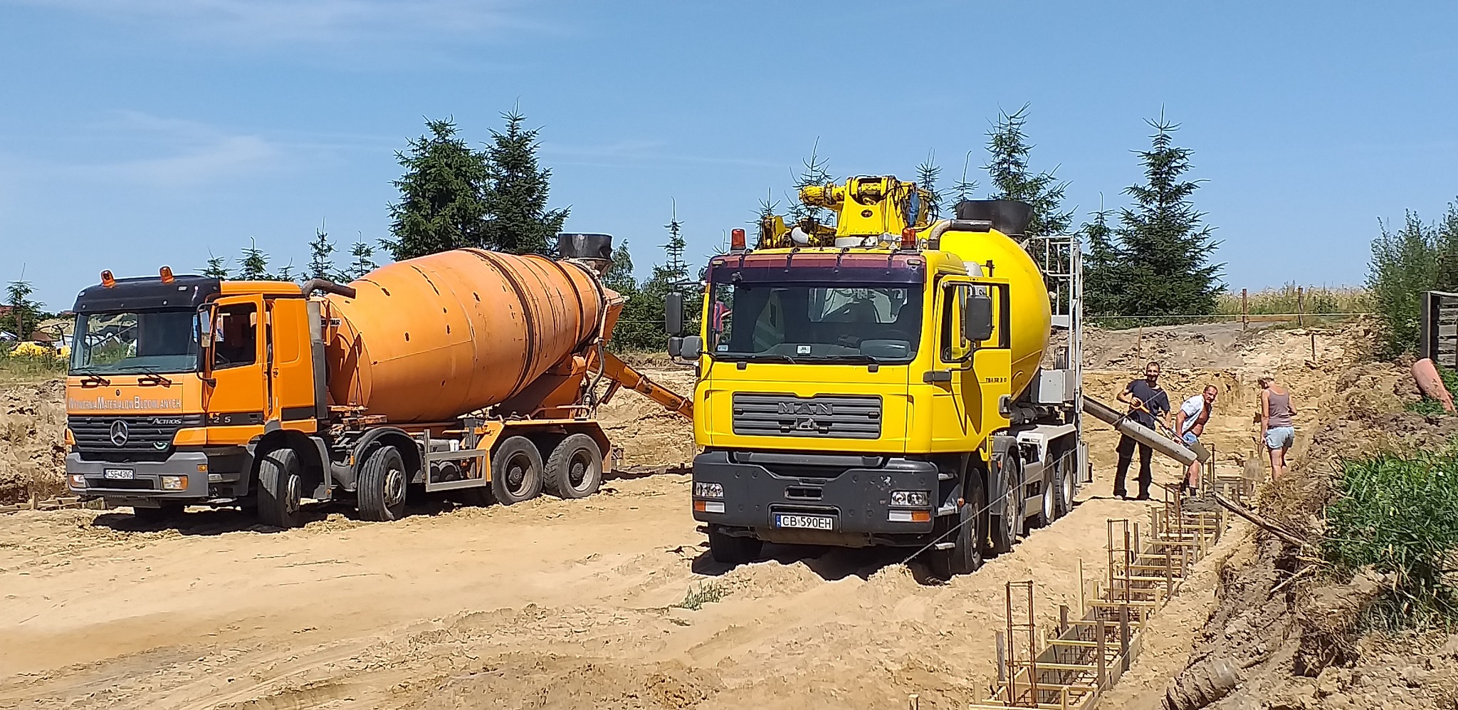 Dwa samochody betonomieszarki, pomarańczowy Mercedes i żółty MAN, stoją na placu budowy obok wykopu z przygotowanym zbrojeniem fundamentów, gdzie robotnicy wylewają beton.